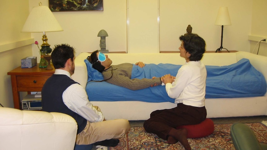 A person wearing headphones and eye mask lies on a couch, attended by two guides to provide support and reassurance.