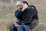 A woman, sitting in a field with a man, wraps her arms around him as he cries. 