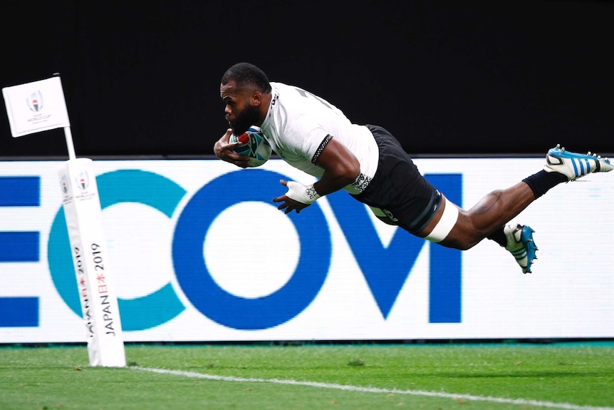 A male rugby union player dives in the air with the ball tucked under his right arm as he scores a try.