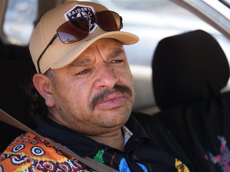 Homem com bigode usando boné dos Raiders e óculos escuros sentado em cima