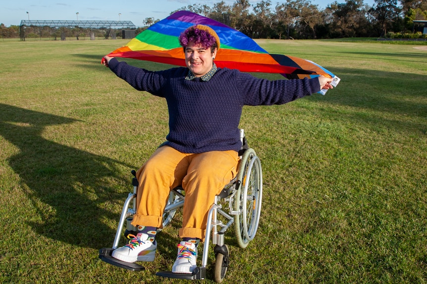 A man in a wheelchair in a park. he holds a pride flag out behind him