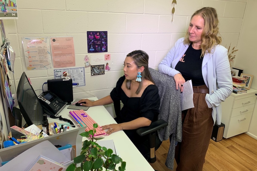 Two women, one standing, look at a computer screen in a small office