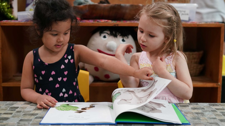 Two young girls sit next to each other at a table looking down at a picture book together.