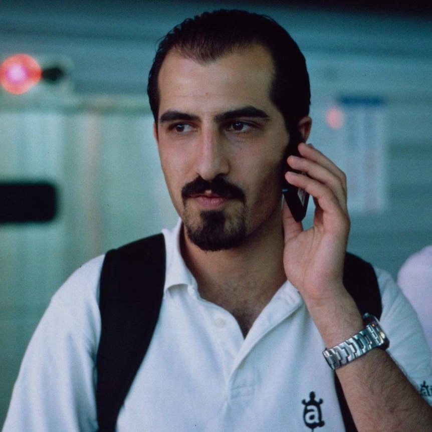Bassel Khatabil Safadi, sporting a cropped beard and wearing a white shirt and black backpack, holds a mobile phone to his ear.
