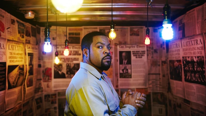 Ice Cube photographed from the side, turning his head to camera. He's in a small room with newspaper on the walls.