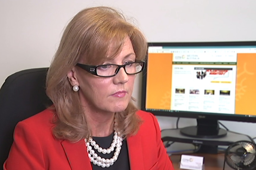 A red-haired woman in a red jacket sits at a desk with a computer screen beside her