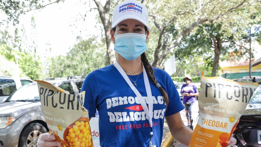A woman in a cap, face mask and blue t-shirt holds up two bags of popcorn to the camera.