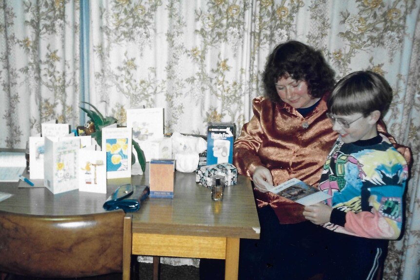 A young boy and his mother, sitting at the table in a 1980s dining room, look at a birthday card.