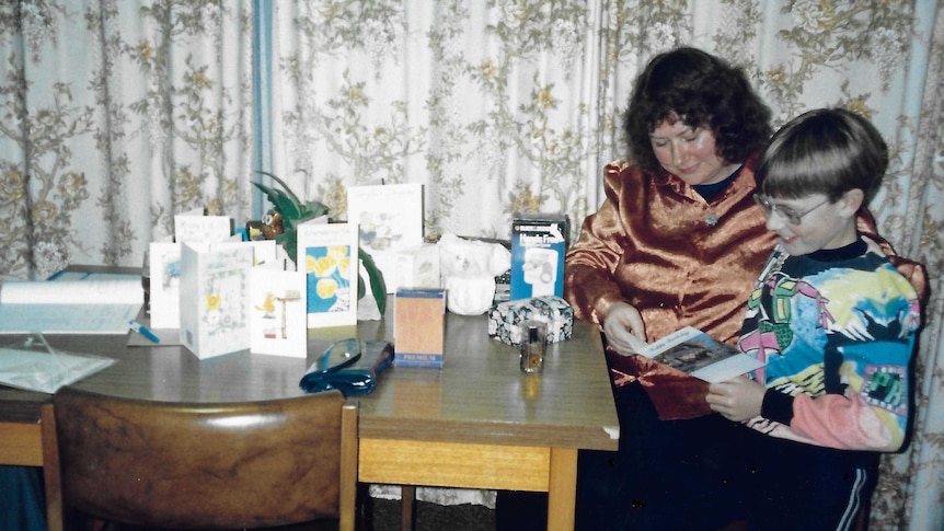 A young boy and his mother, sitting at the table in a 1980s dining room, look at a birthday card.