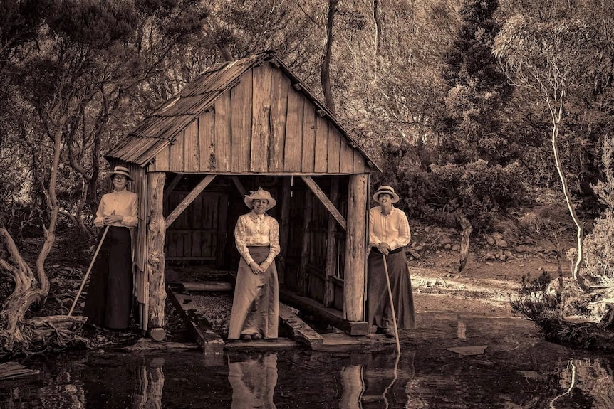 Three women in Edwardian dress standing near and old fashioned timber boatshed.