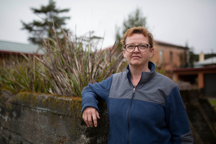 A woman wearing glasses leans against a stone wall outside and looks at the camera.