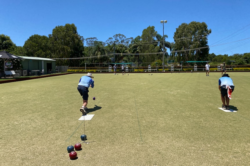 Two men bend over and bowl on a bowling green whilst other men look at them from the other end of the green