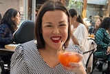 Woman grins in restaurant, raising a cocktail to the camera