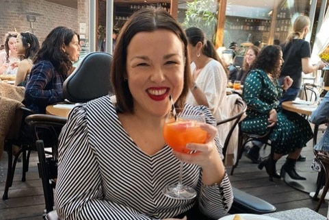Woman grins in restaurant, raising a cocktail to the camera