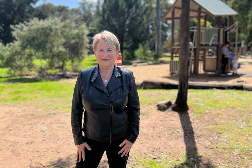 A woman with short, blonde hair, wearing a dark outfit, stands near a playground.