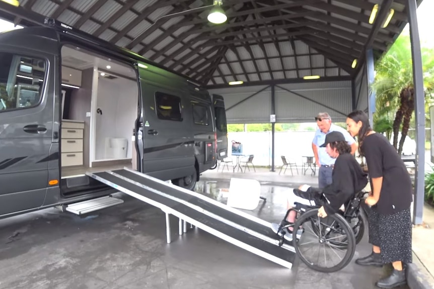 A man on a wheelchair with a woman pushing him up a ramp into a large campervan.