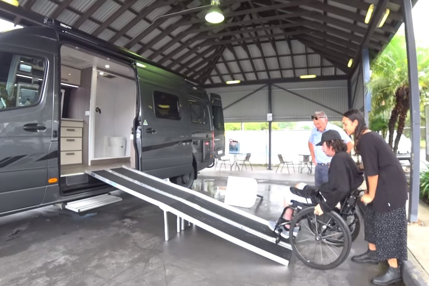 A man in a wheelchair with a woman pushing him up a ramp into a large motor home.