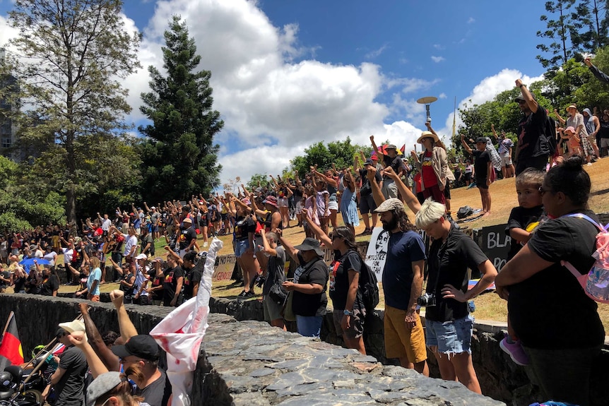 Hundreds of people stand on a natural amphitheatre, each raising one fist in the air.