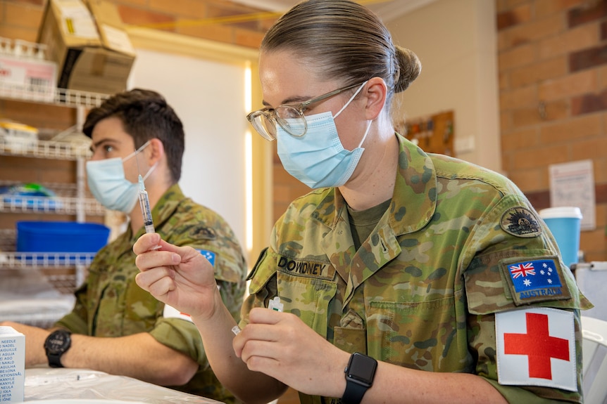 A woman wearing an Australian Army Uniform and a facemask prepares a syringe.