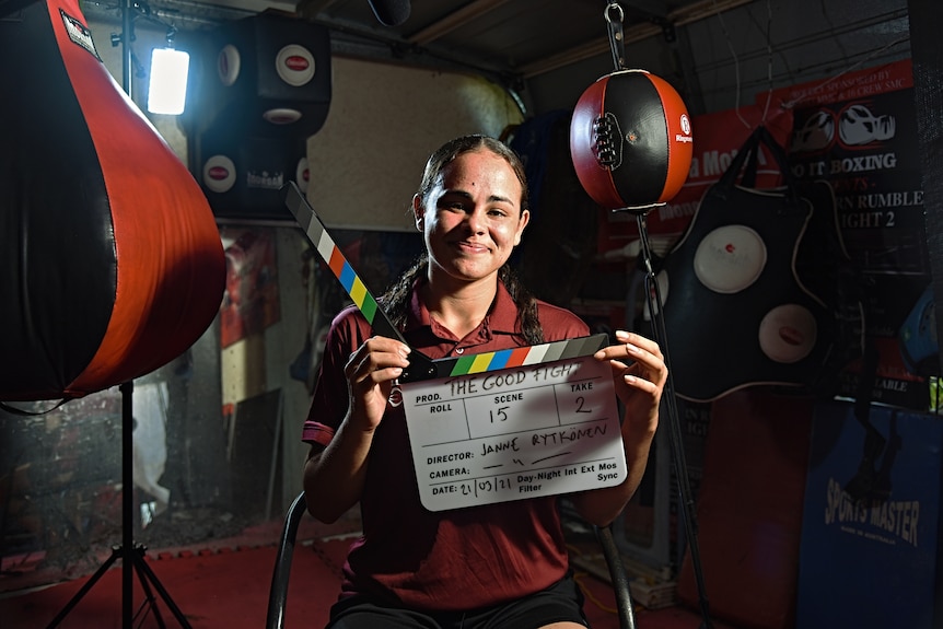 Boxer holds up film slate/clapper board in gym surrounded by boxing equipment.