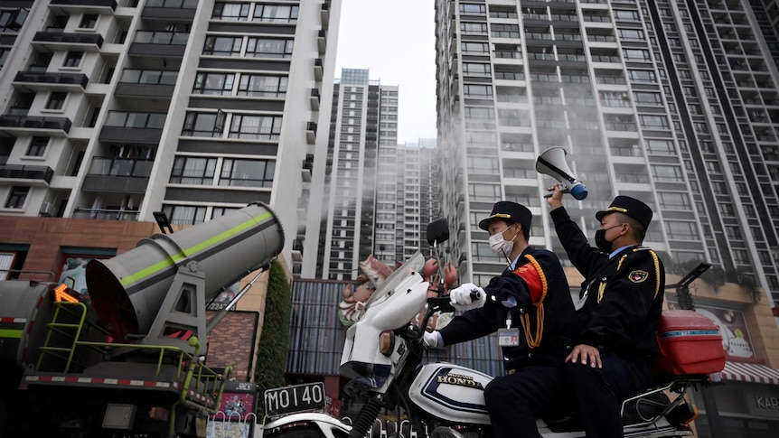 From a low angle you look up at two police officers on a motorcycle as it drives past a sanitizing vehicle spouting disinfectant