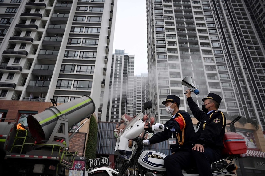 From a low angle you look up at two police officers on a motorcycle as it drives past a sanitizing vehicle spouting disinfectant