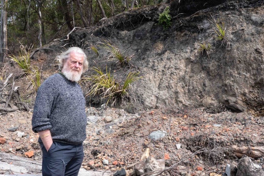 An older man with a white beard stands near an eroded cliff