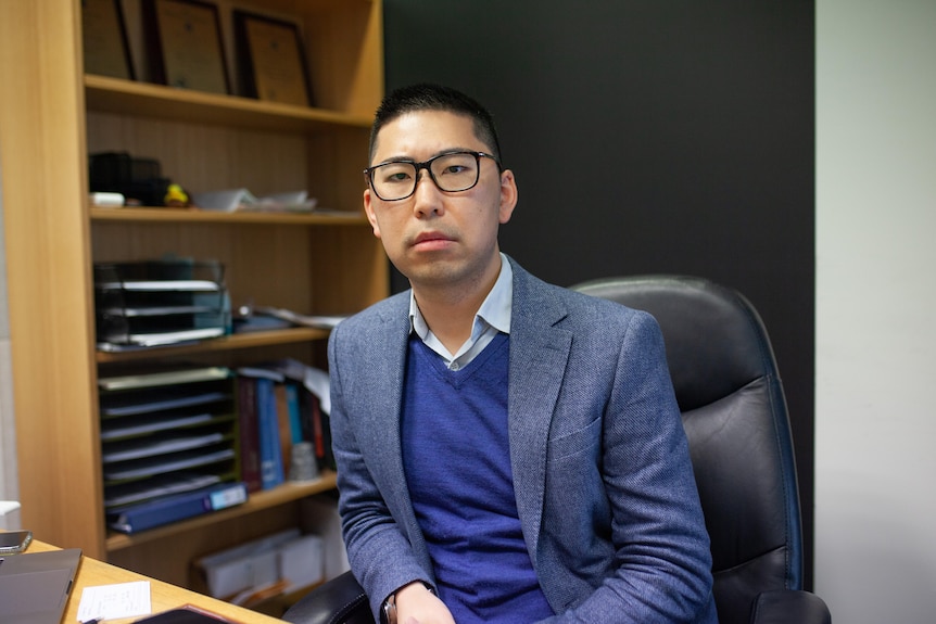 A man in a suit and glasses sits at a desk, looking at the camera.