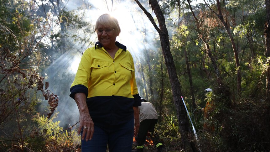 An older Indigenous woman wearing high-vis stands smiling a dense forest.