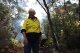 An older Indigenous woman wearing high-vis stands smiling a dense forest.