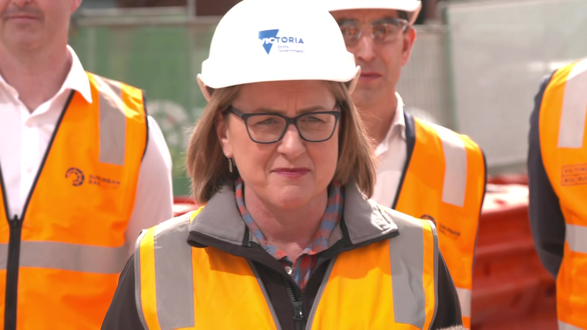 Jacinta Allan wears a white hard hat with the Victorian government logo and an orange high vis vest and stands with others.