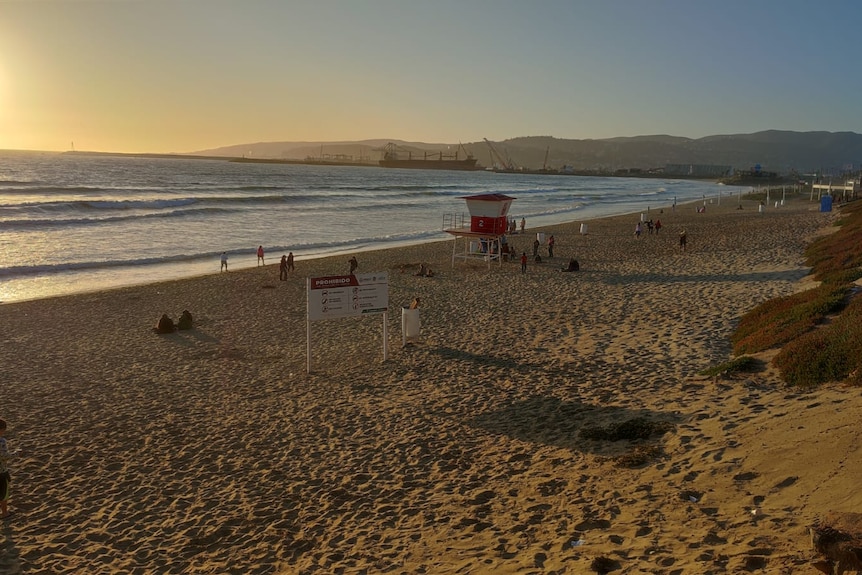 A wide shot of a sandy beach with a few people standing watching the sunset.