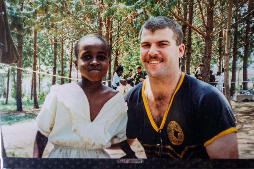 A male volunteer smiles at the camera, he has his arm around a young Rwandan child.
