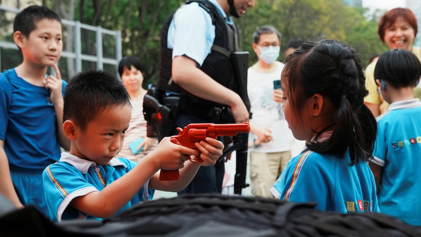 A boy points a red plastic pistol toy in the face of a girl standing less than a metre away as smiling adults and police watch.