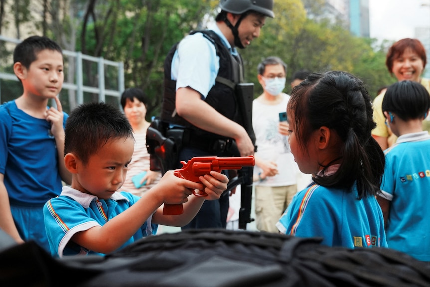 A boy points a red plastic pistol toy in the face of a girl standing less than a metre away as smiling adults and police watch.