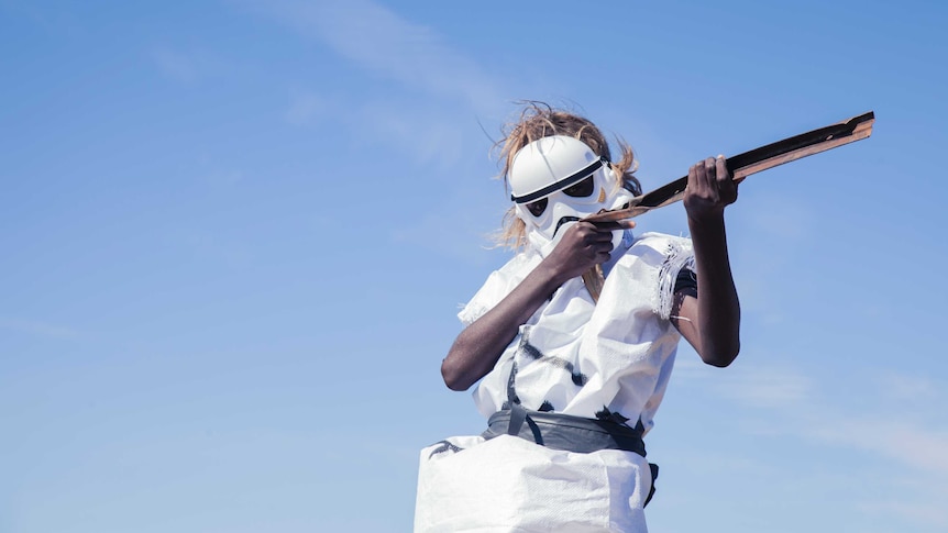 A young girl stands against a stark blue sky, wearing a plastic storm-trooper mask and holding a makeshift blaster.