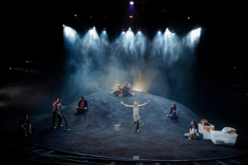 On a dimly lit stage, Indigenous people are arranged on a mound, including a cultural dancer, and a man with a didgeridoo