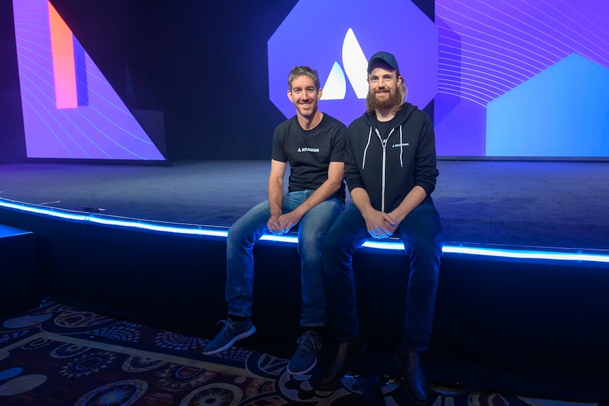 Two men sit on a stage wearing t-shirts and jeans.