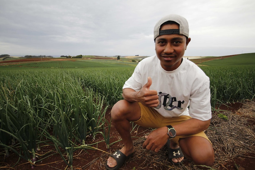 A young man grins and gives the thumbs up as he squats next to plants growing in a field.