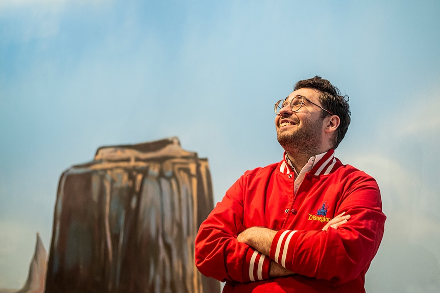 Colour photograph of artist David Capra standing in front of a painted Monument Valley backdrop, looking up towards the ceiling.