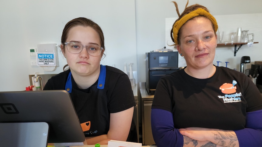 Two people wearing shirts that say "a kinder cup cafe" look unhappy behind a counter bench