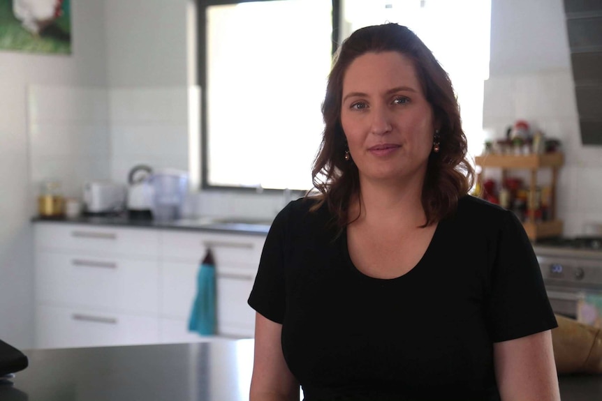 A lady in a black tshirt standing in a kitchen with lots of sunlight coming through the window