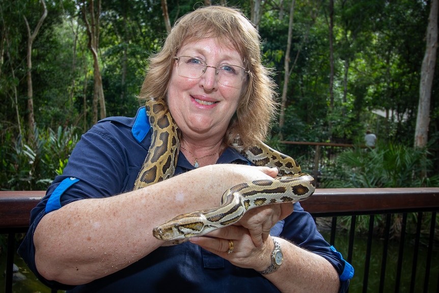 A smiling, bespectacled middle-aged woman with fair hair stands in a nature park, holding a python.