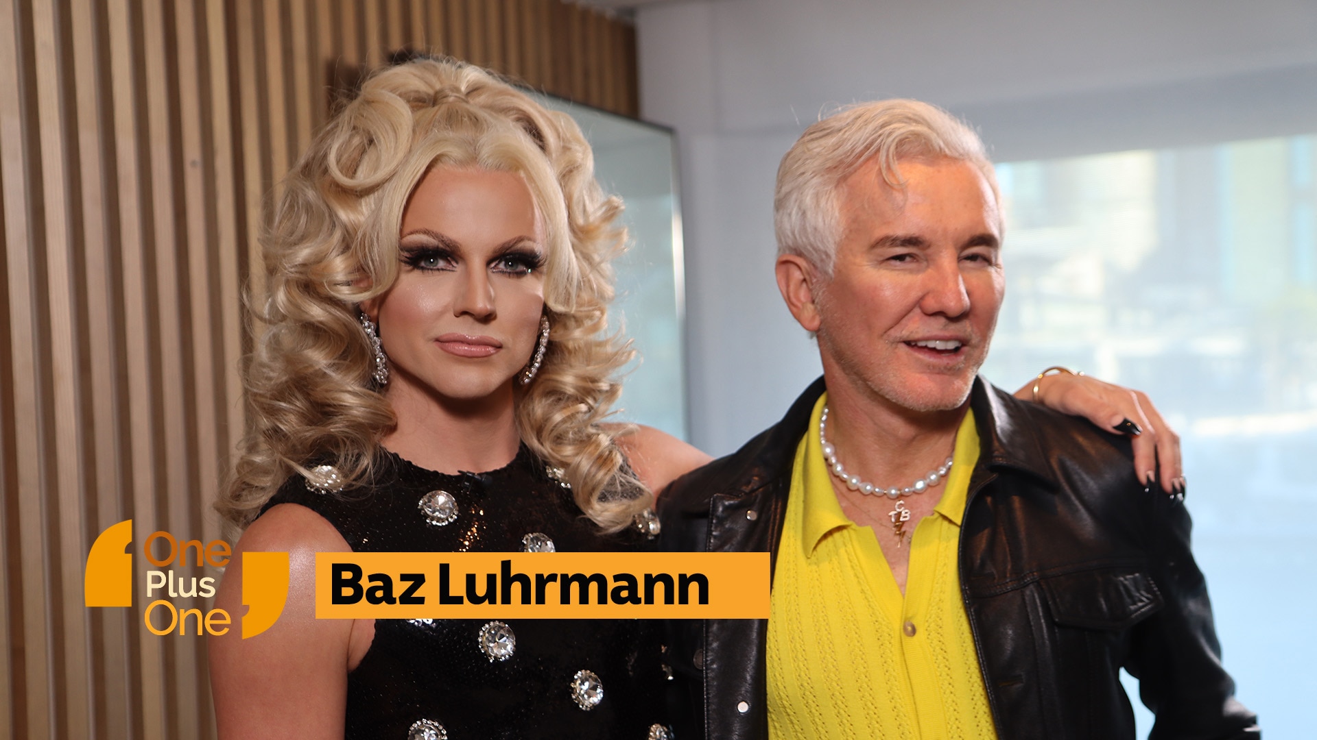 Glamourously dressed drag queen with blonde curly hair stands next to white-haired man with yellow shirt and leather jacket