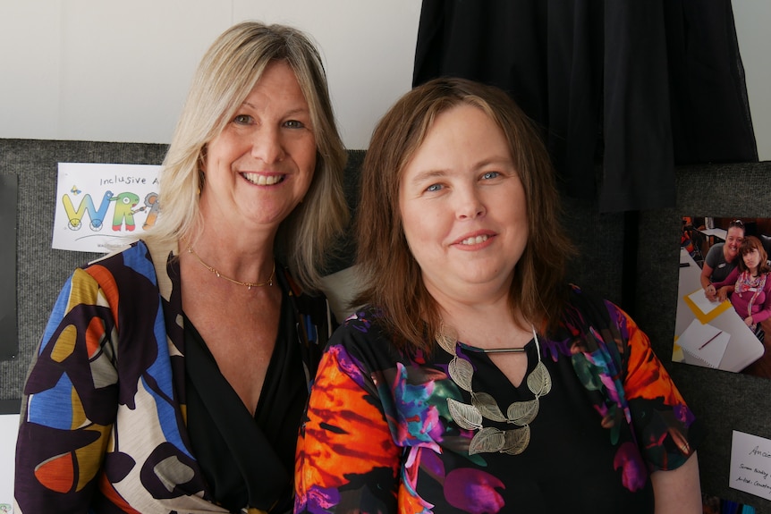 Two women in floral shirts stand side by side and smile with artwork in the background 