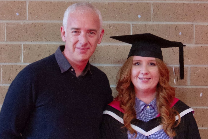 A young woman in a graduation gown with an older man in a jumper posing for a photo
