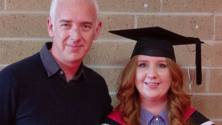 A young woman in a graduation gown with an older man in a jumper posing for a photo