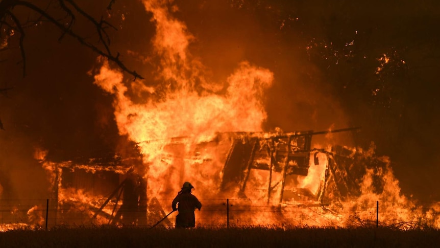 A firefighter is seen in front of a large bushfire burning through a structure.