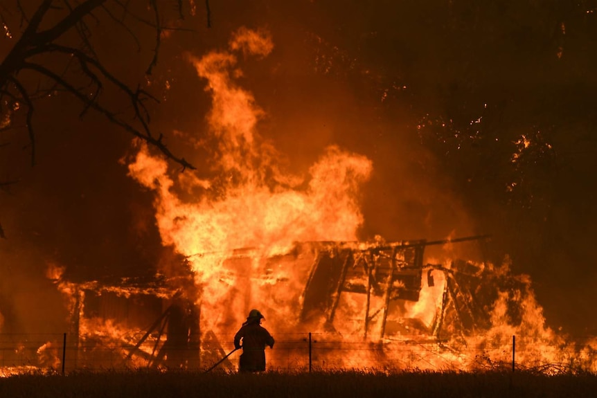 A firefighter is seen in front of a large bushfire burning through a structure.