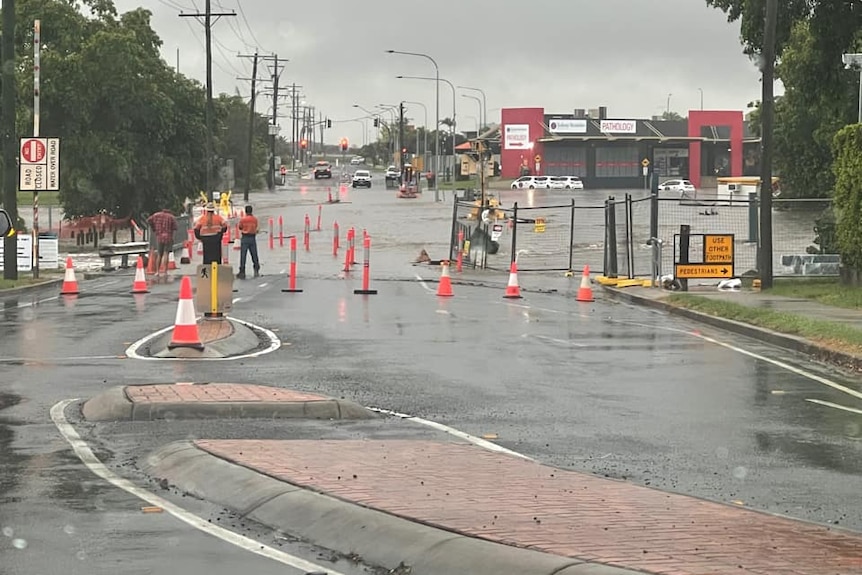 a road with water over it in a residential and commercial area, safety signs are out and road workers about